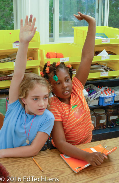 Two third-grade girls raise hands to answer teacher’s questions